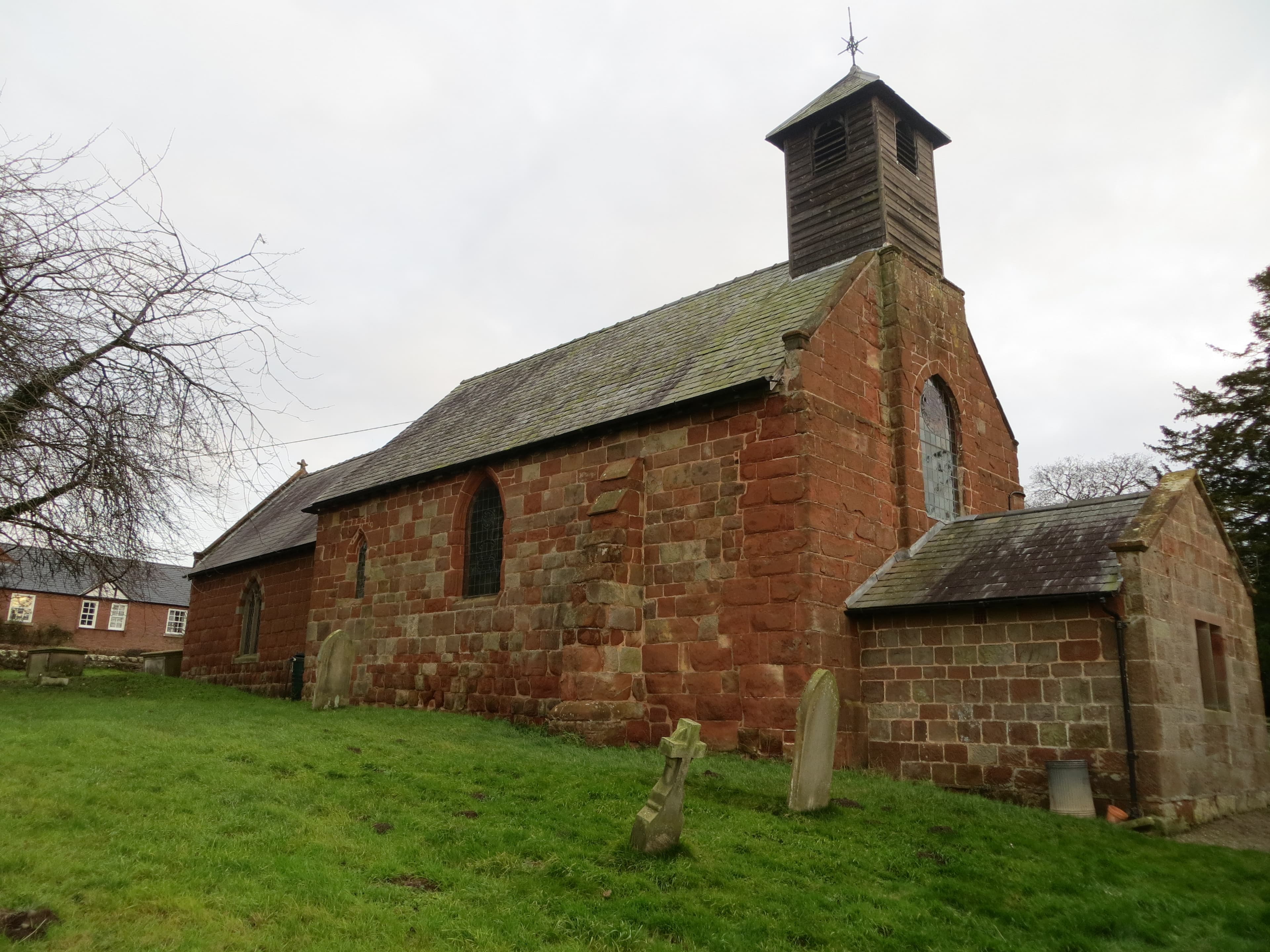 Red sandstone church with a timber bellcote and pitched roofs, with gravestones in the grass in front.